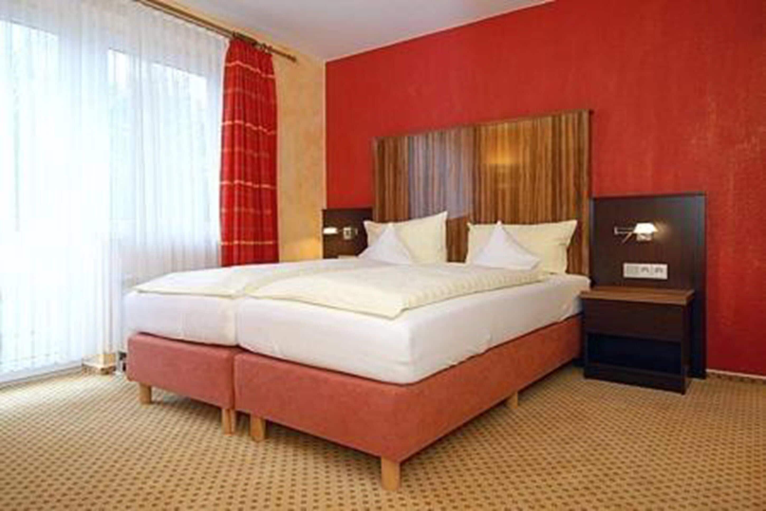 Cama doble con cabecera de madera, dos mesitas de noche con lámparas y una pared de acento roja.