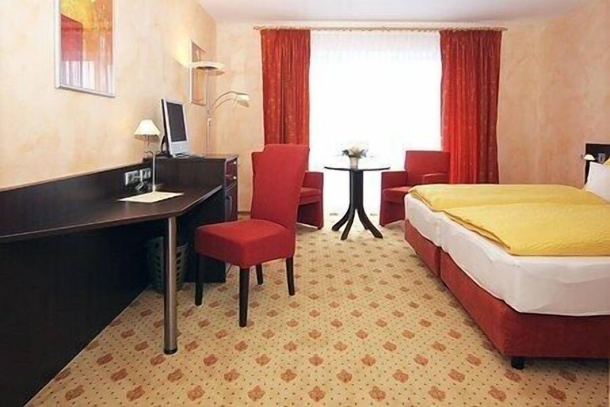 Una habitación de hotel con cama, escritorio, silla, mesita y ventana con cortinas.