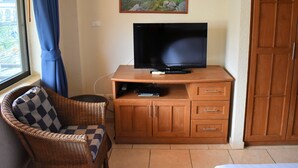 A room with a television on a wooden cabinet, a wicker chair with a checkered cushion, and a painting of a coastal landscape on the wall. - Klein Heidelberg (Pattaya)
