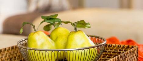 A bowl of green pears on a woven mat with a wicker basket underneath, placed on a bed with a white pillow and a lamp in the background.