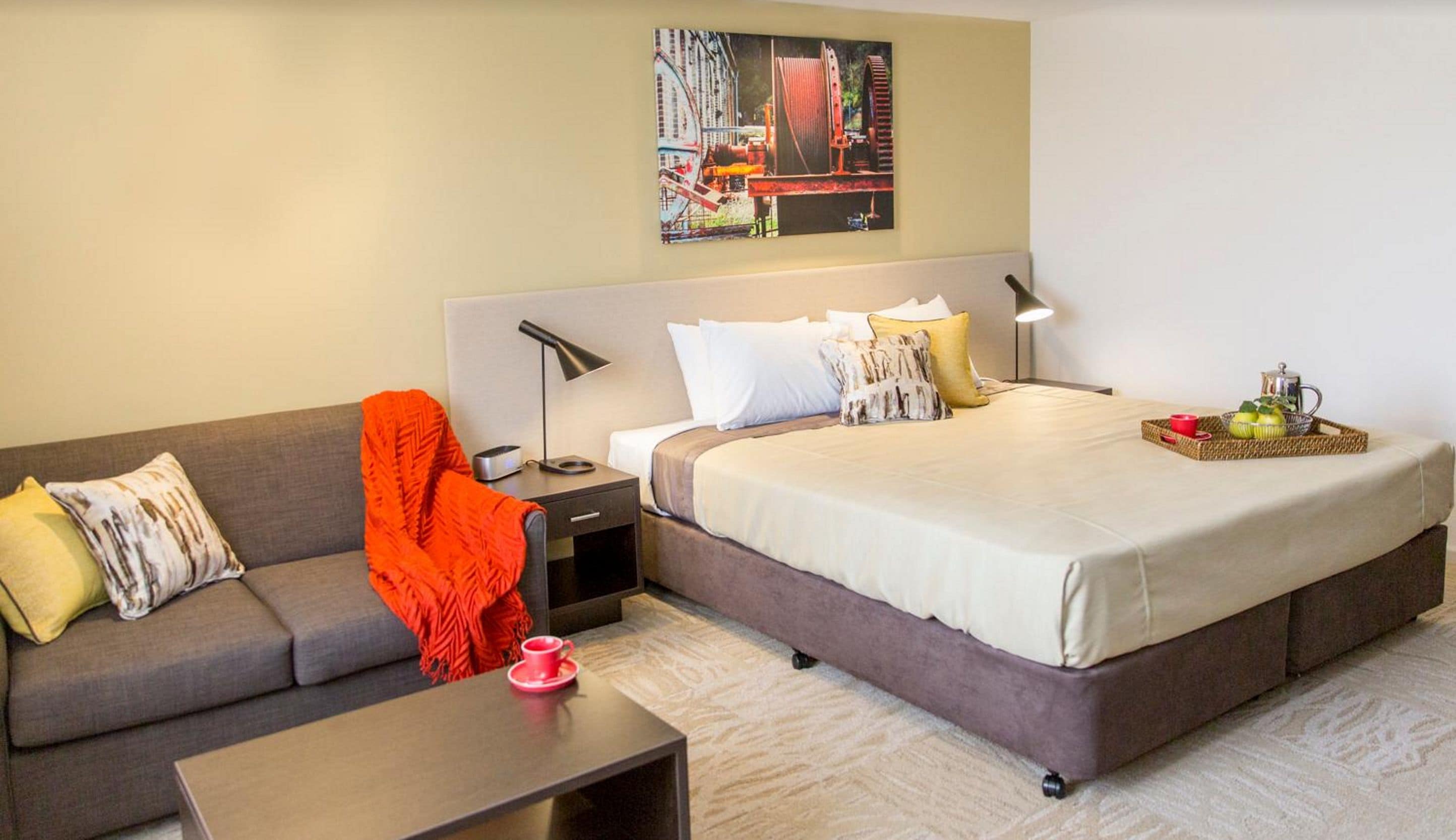 A hotel room with a bed, a sofa, a nightstand, a lamp, and a painting on the wall.
