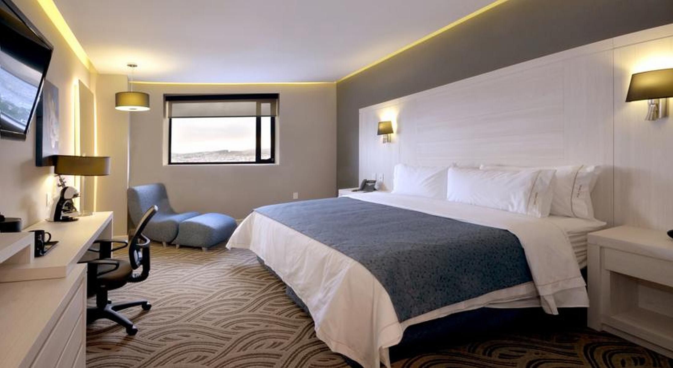Ein Hotelzimmer mit Bett, Schreibtisch, Stuhl und einem an der Wand befestigten Fernseher.