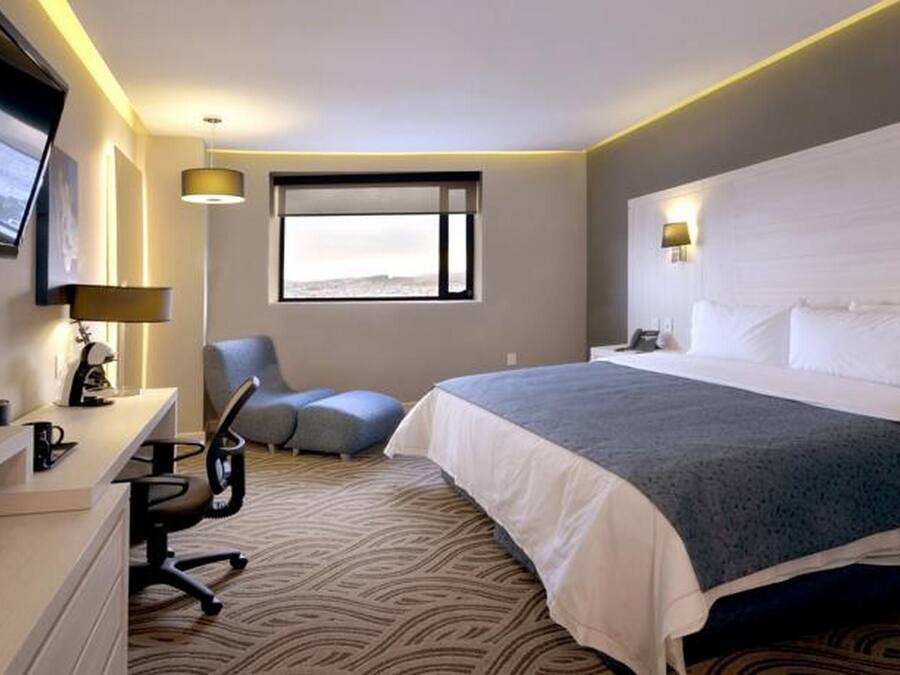 Ein Hotelzimmer mit Bett, Schreibtisch, Stuhl und einem an der Wand befestigten Fernseher.