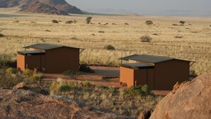 Two small, modern, brown-roofed structures in a dry, open landscape with sparse vegetation and distant mountains.