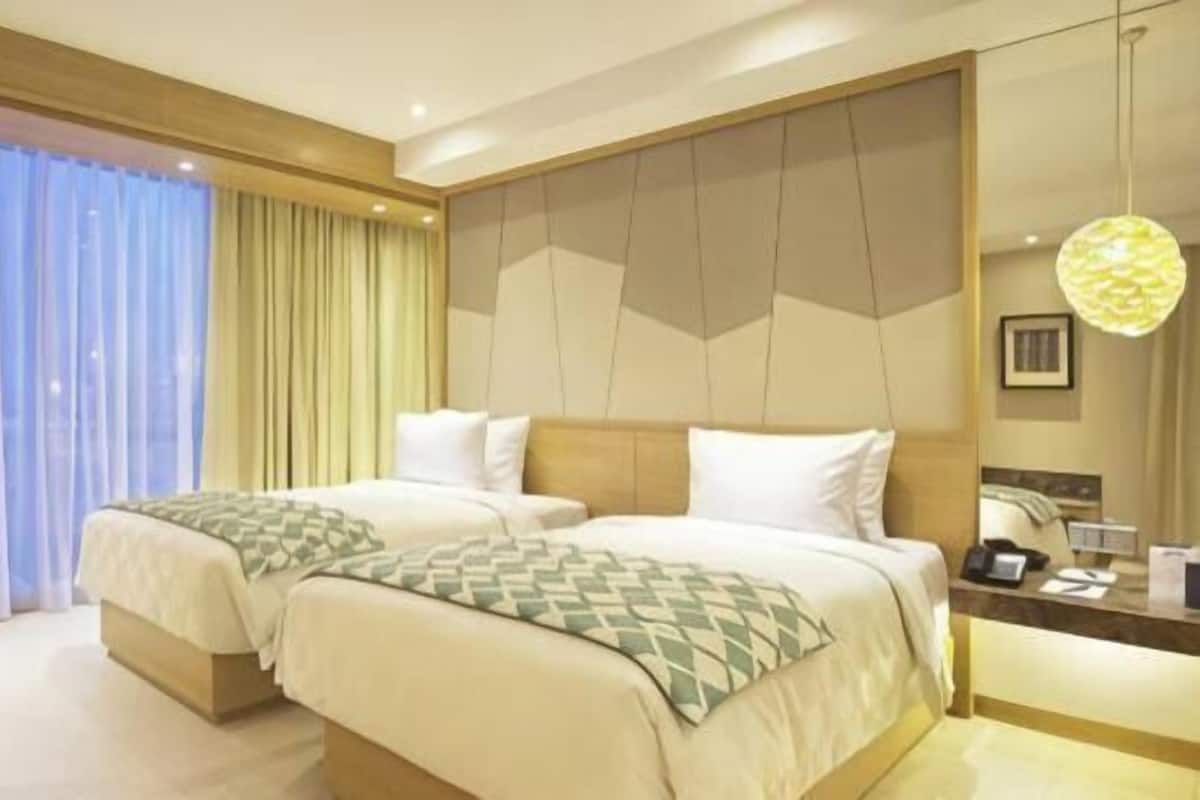 Une chambre d’hôtel avec deux lits, une grande fenêtre avec des rideaux, une tête de lit en bois et une table de chevet avec une lampe.