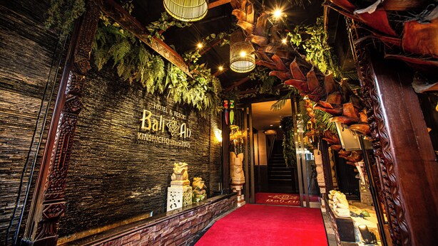 L’entrée d’un hôtel, avec un tapis rouge, un mur en bois portant le nom de l’hôtel, ainsi que des plantes et des sculptures décoratives.