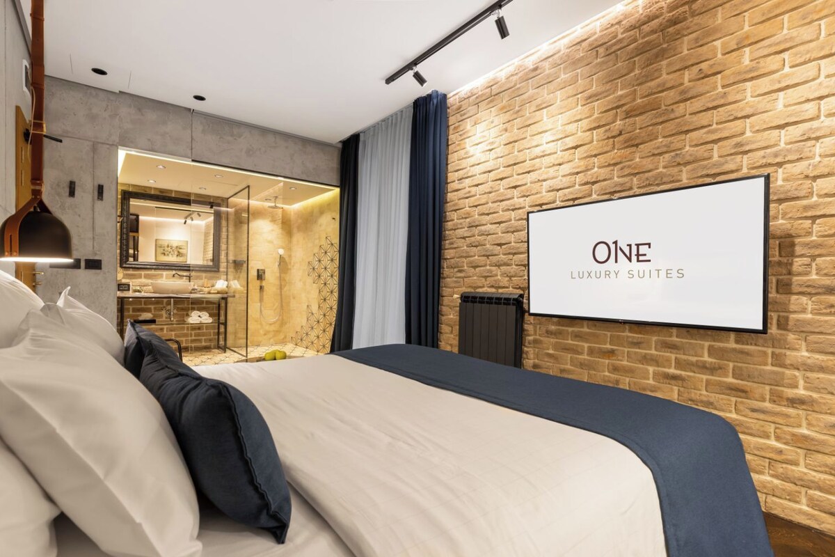 Una habitación de hotel moderna con una cama grande, un baño con paredes de vidrio y una pared de ladrillo.