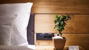 Una cama bien hecha con sábanas blancas, una pequeña planta en maceta sobre una mesita de noche de madera y un cartel que indica acceso a WiFi gratuito.