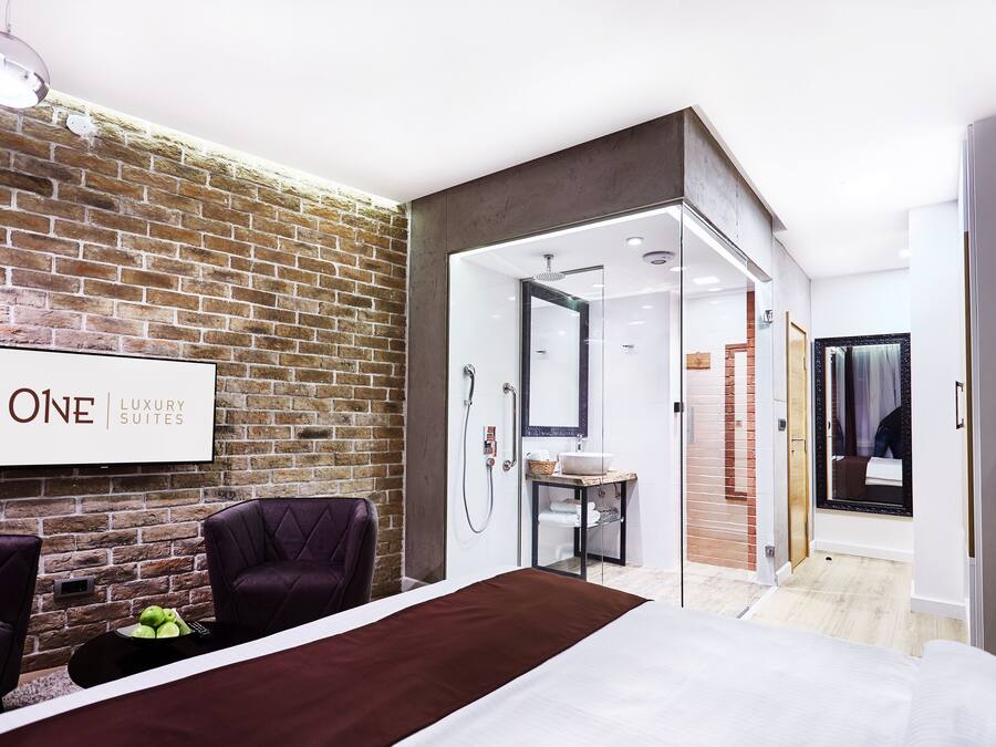 Habitación de hotel moderna con pared de ladrillo, cama, zona de estar con mesa y baño visible a través de una puerta abierta.