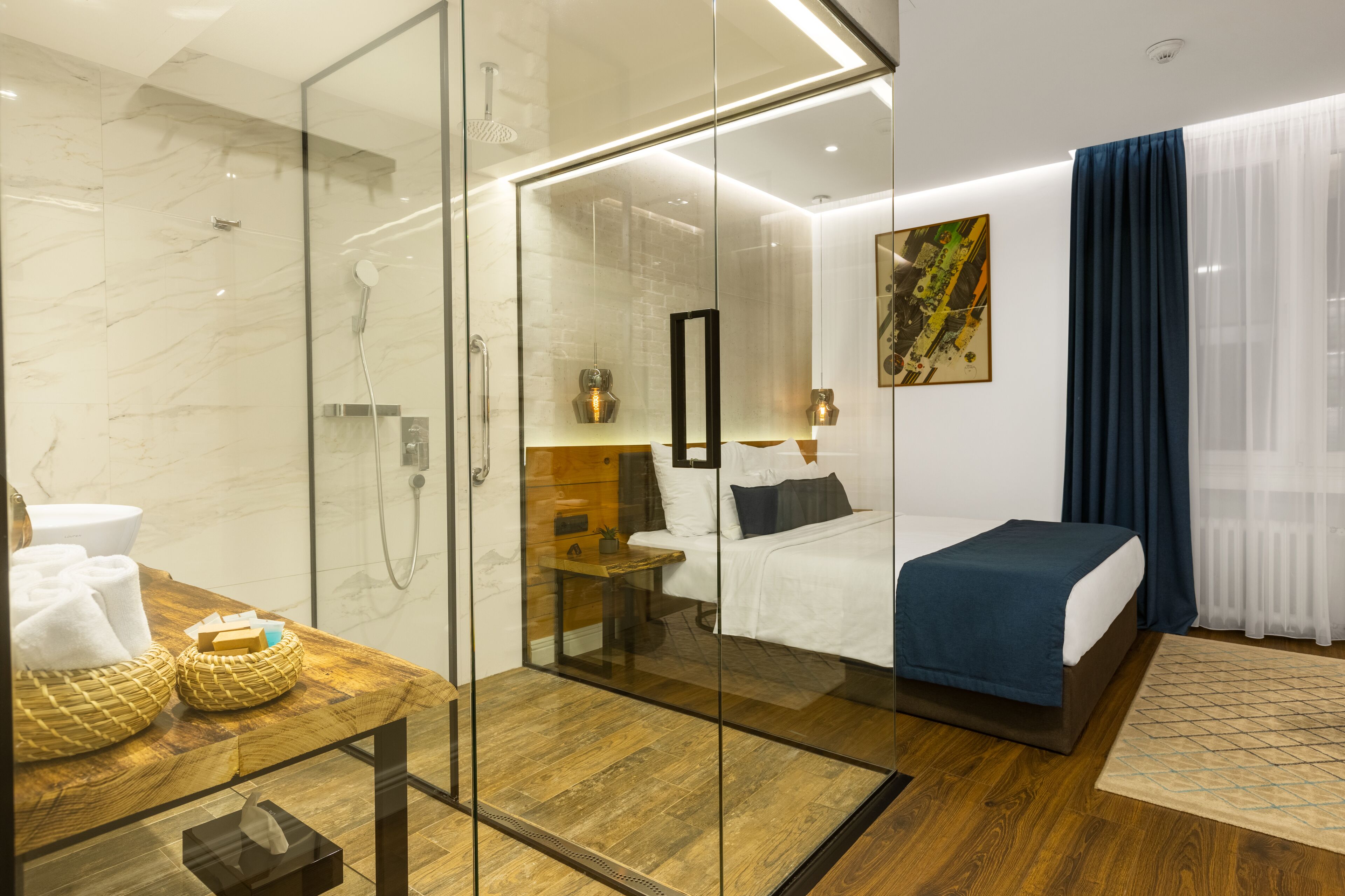 Une chambre d’hôtel moderne dotée d’une douche en verre, d’une coiffeuse en bois, d’un lit recouvert d’une courtepointe bleue et d’un grand tableau abstrait accroché au mur.