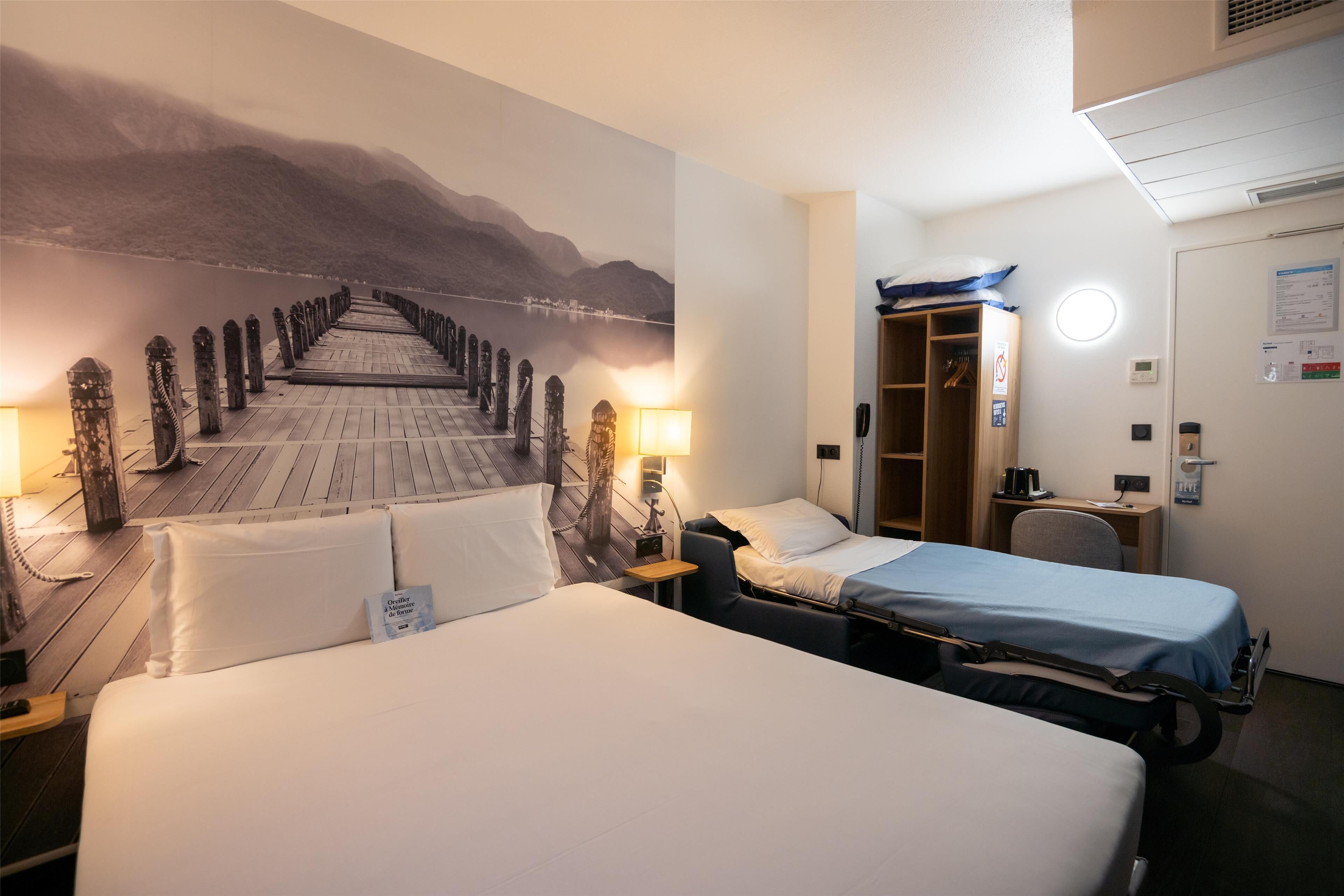 Een hotelkamer met twee bedden, een bureau, een stoel, een kledingkast en een muurschildering van een brug.