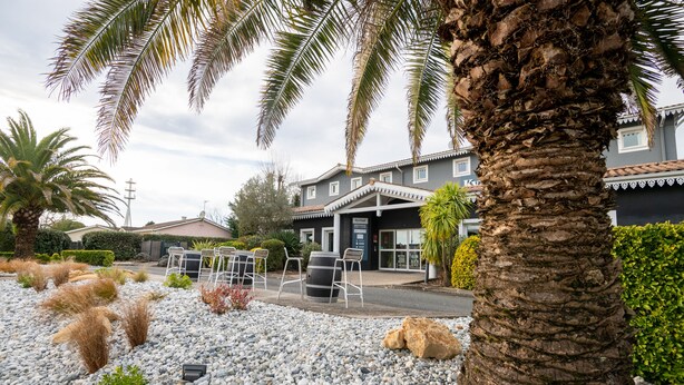 A building with a covered outdoor seating area, surrounded by palm trees and a gravel ground. - KYRIAD BORDEAUX SUD - Villenave d'Ornon (Villenave-d'Ornon)