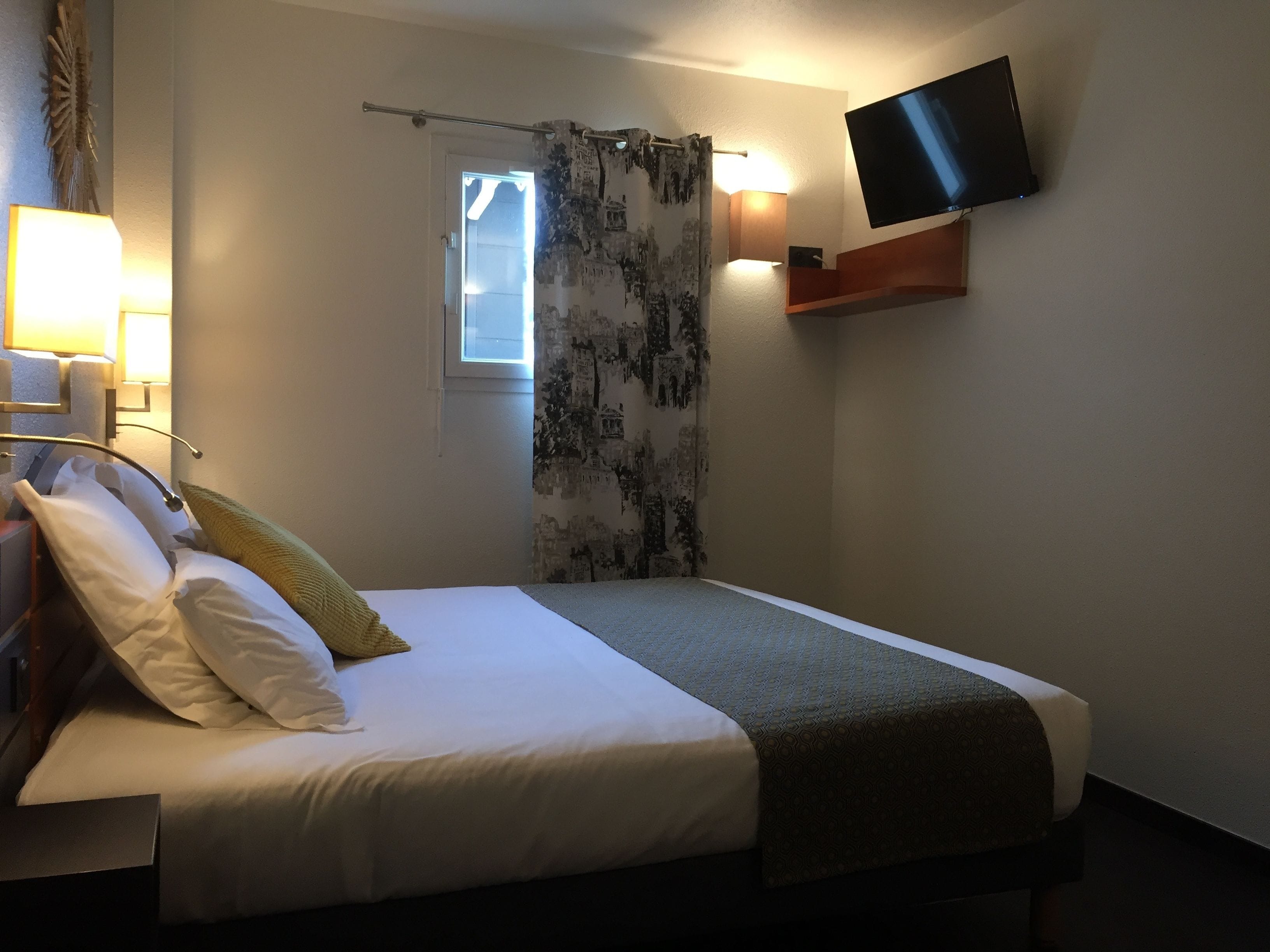 A neatly made bed with white linens and a patterned throw blanket, a bedside lamp, and a flat-screen TV mounted on the wall.