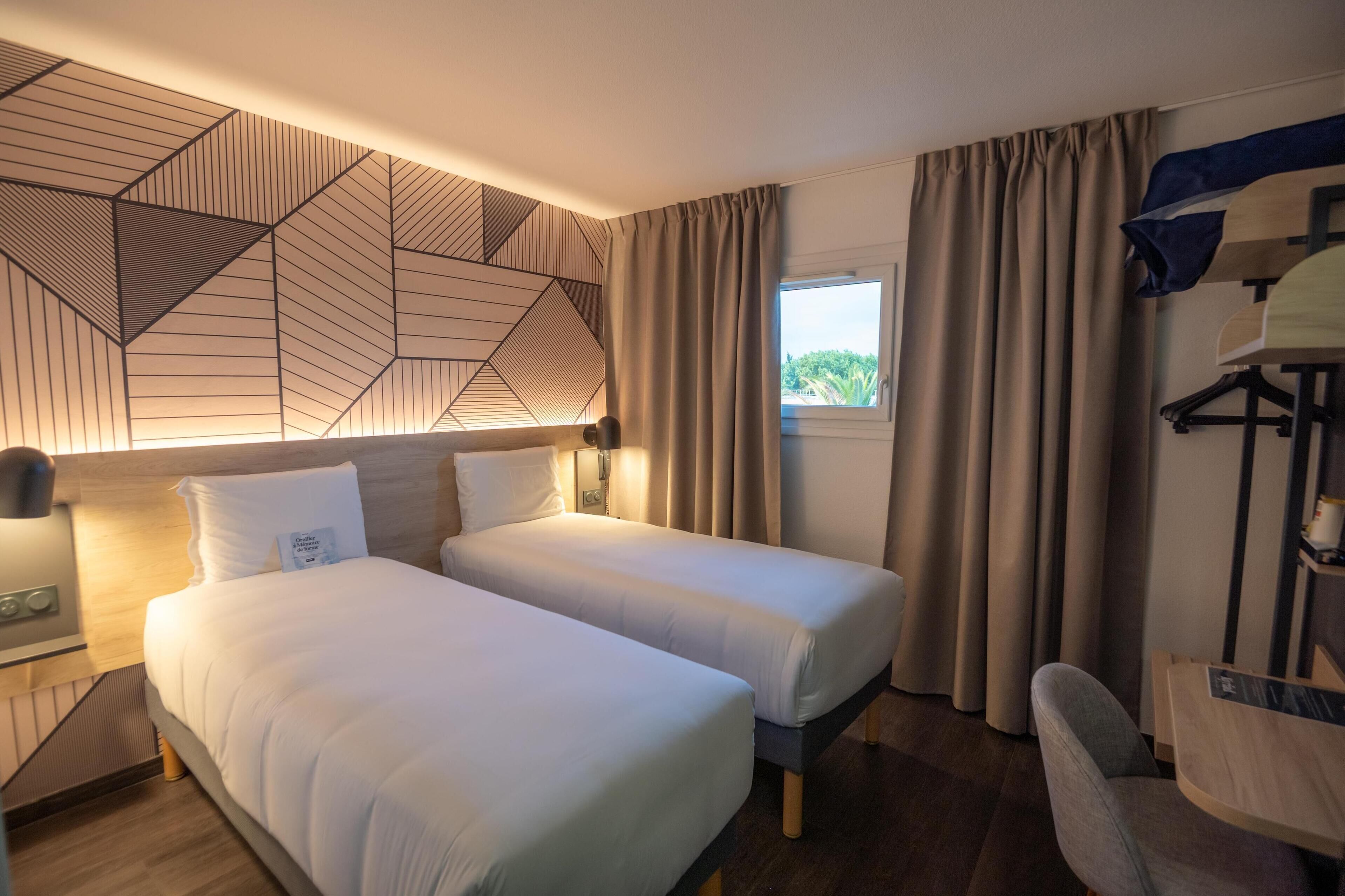 Habitación de hotel con dos camas, un escritorio, una silla y una ventana con cortinas.