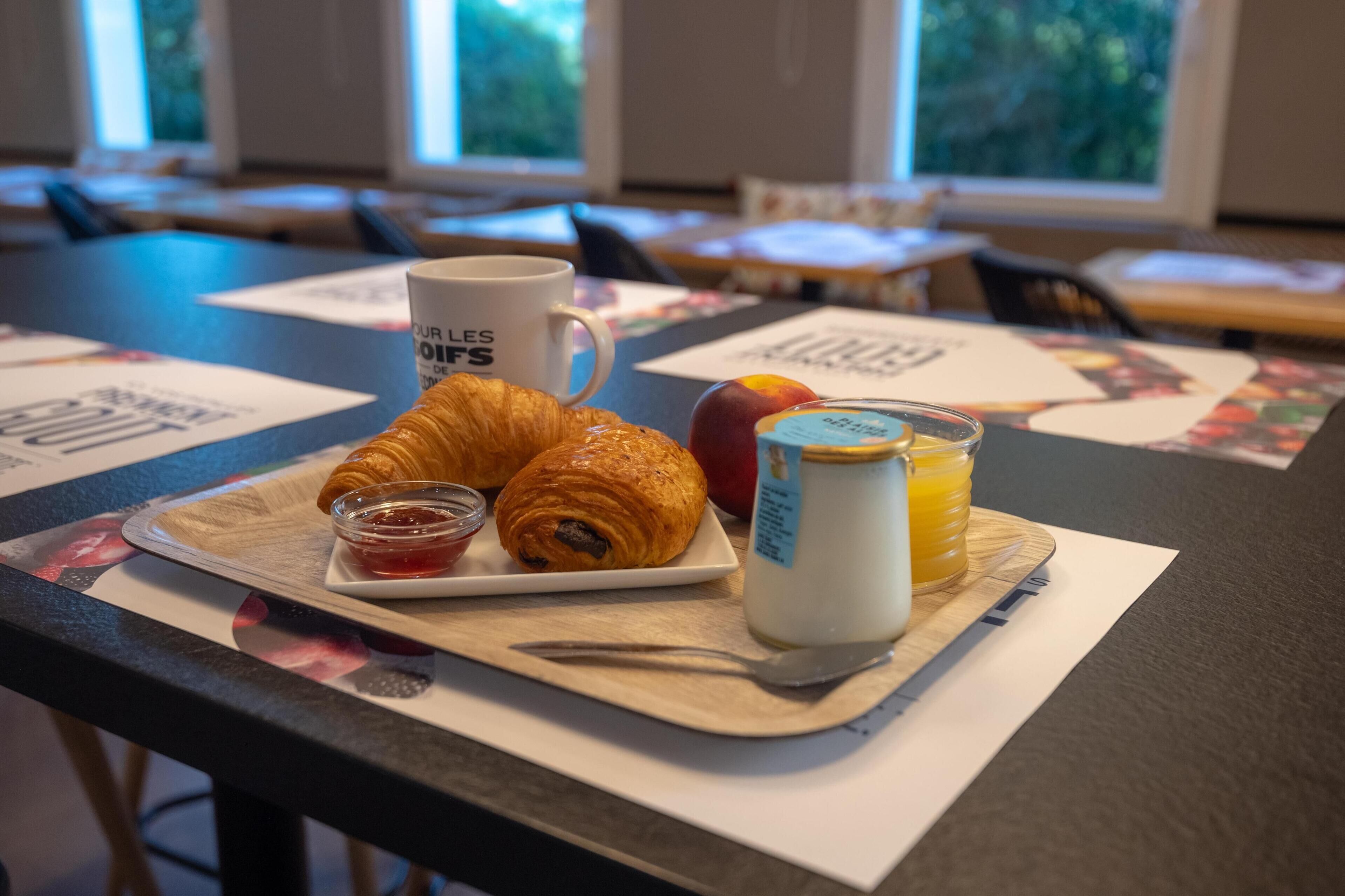 Bandeja de desayuno con cruasanes, mermelada, yogur y fruta sobre una mesa en una habitación con ventanas amplias y vistas a árboles.