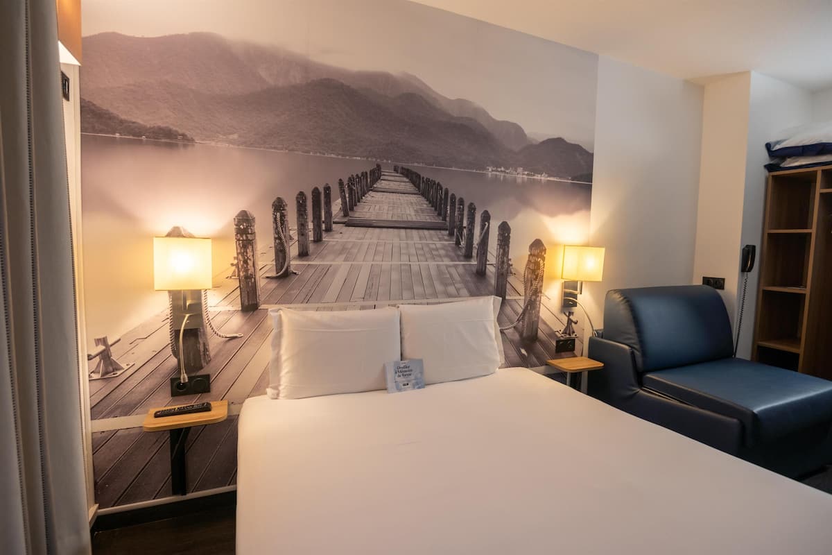 A hotel room with a large bed, two bedside lamps, a blue chair, a wooden desk, and a wall mural of a pier leading to mountains.