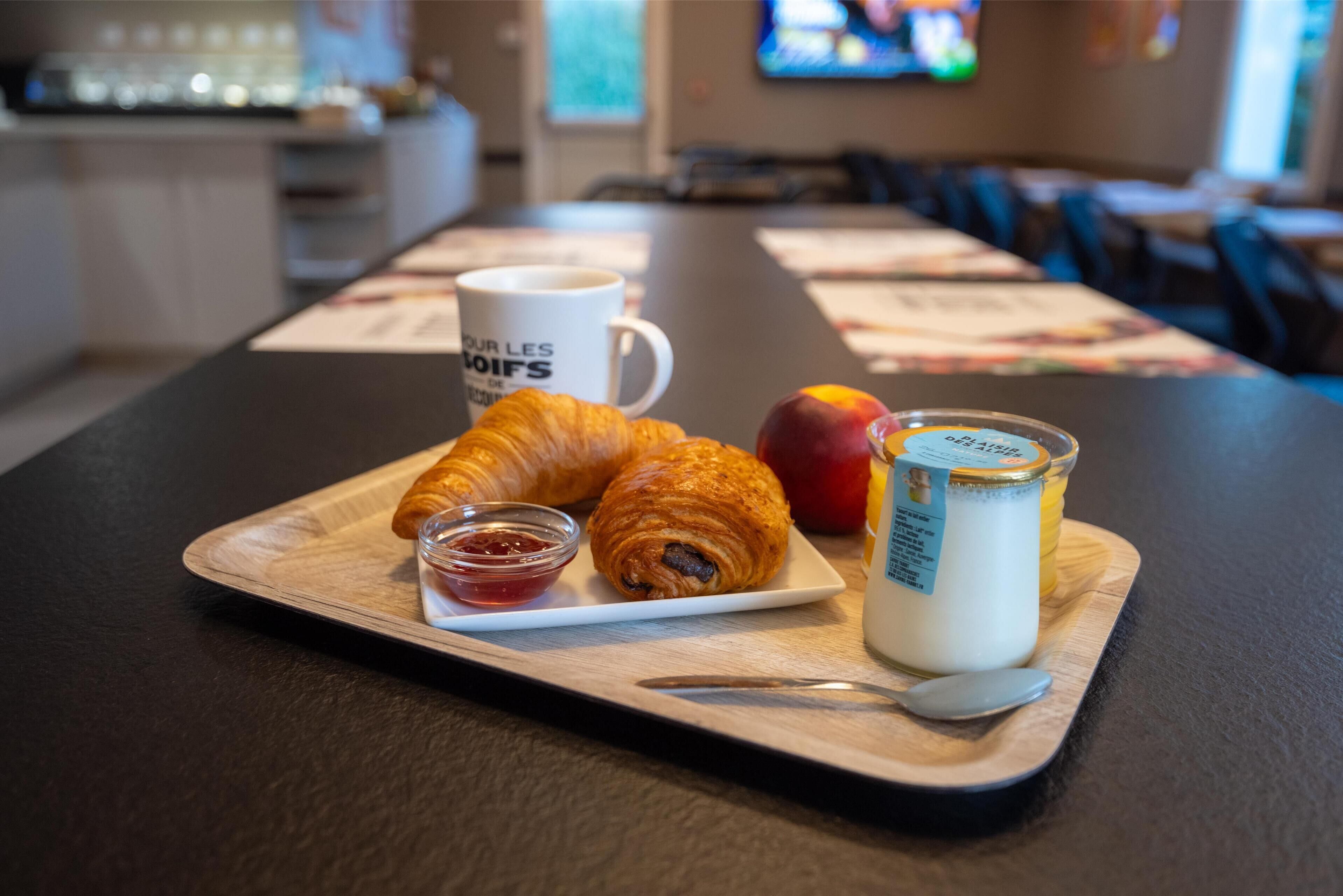 A breakfast tray with croissants, a small container of jam, a glass of milk, and an apple on a wooden board.