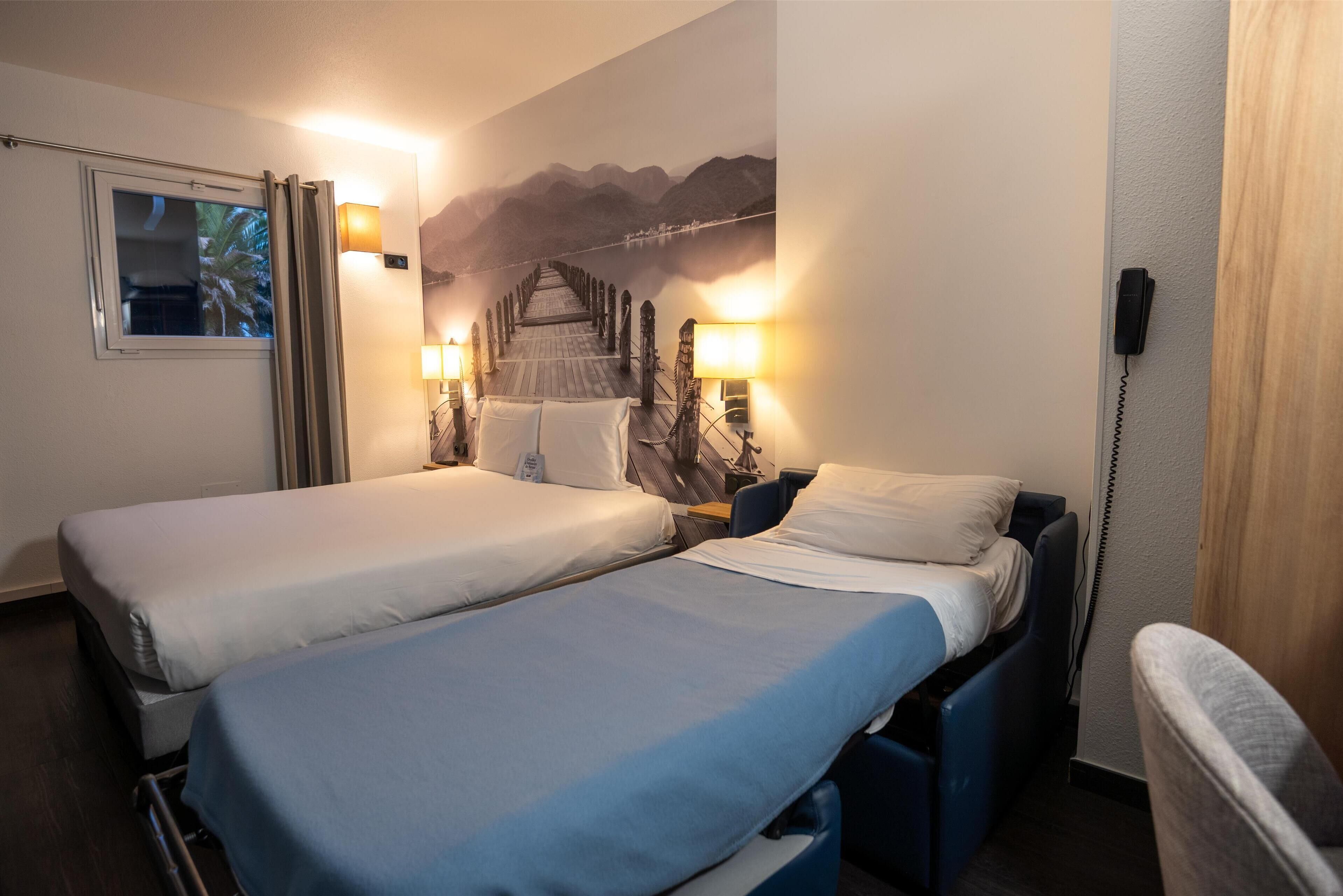 Ein Hotelzimmer mit zwei Einzelbetten, einem Holztisch, einem Stuhl, einem Fenster mit Vorhängen, einem Wandtelefon und einem Wandbild von Bergen.