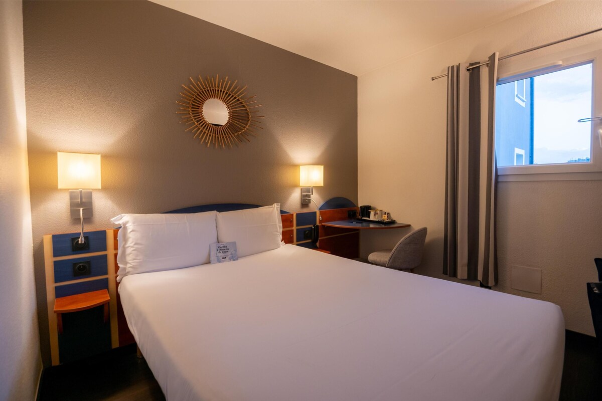 A hotel room with a bed, bedside lamps, a desk, a chair, a window with curtains, and a wall-mounted mirror.