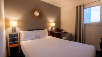 Une chambre d’hôtel comprenant un lit, des lampes de chevet, un bureau, une chaise, une fenêtre avec des rideaux et un miroir fixé au mur.