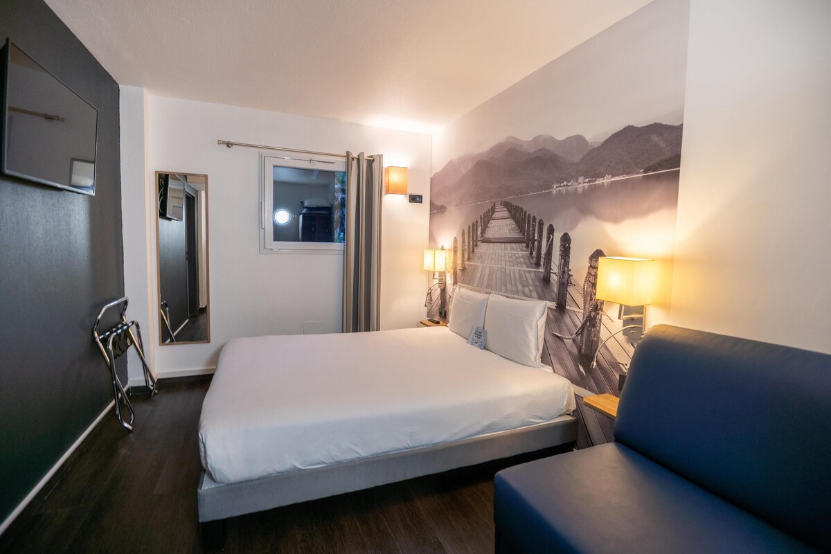 A hotel room with a bed, a blue sofa, a wall-mounted TV, a window, and a painting of a bridge.