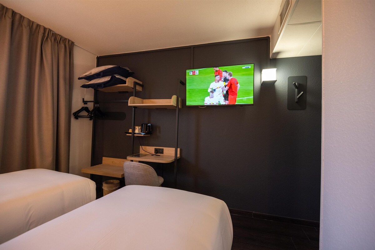 A hotel room with two beds, a TV mounted on the wall, a small table with a chair, and a coat rack with a jacket.