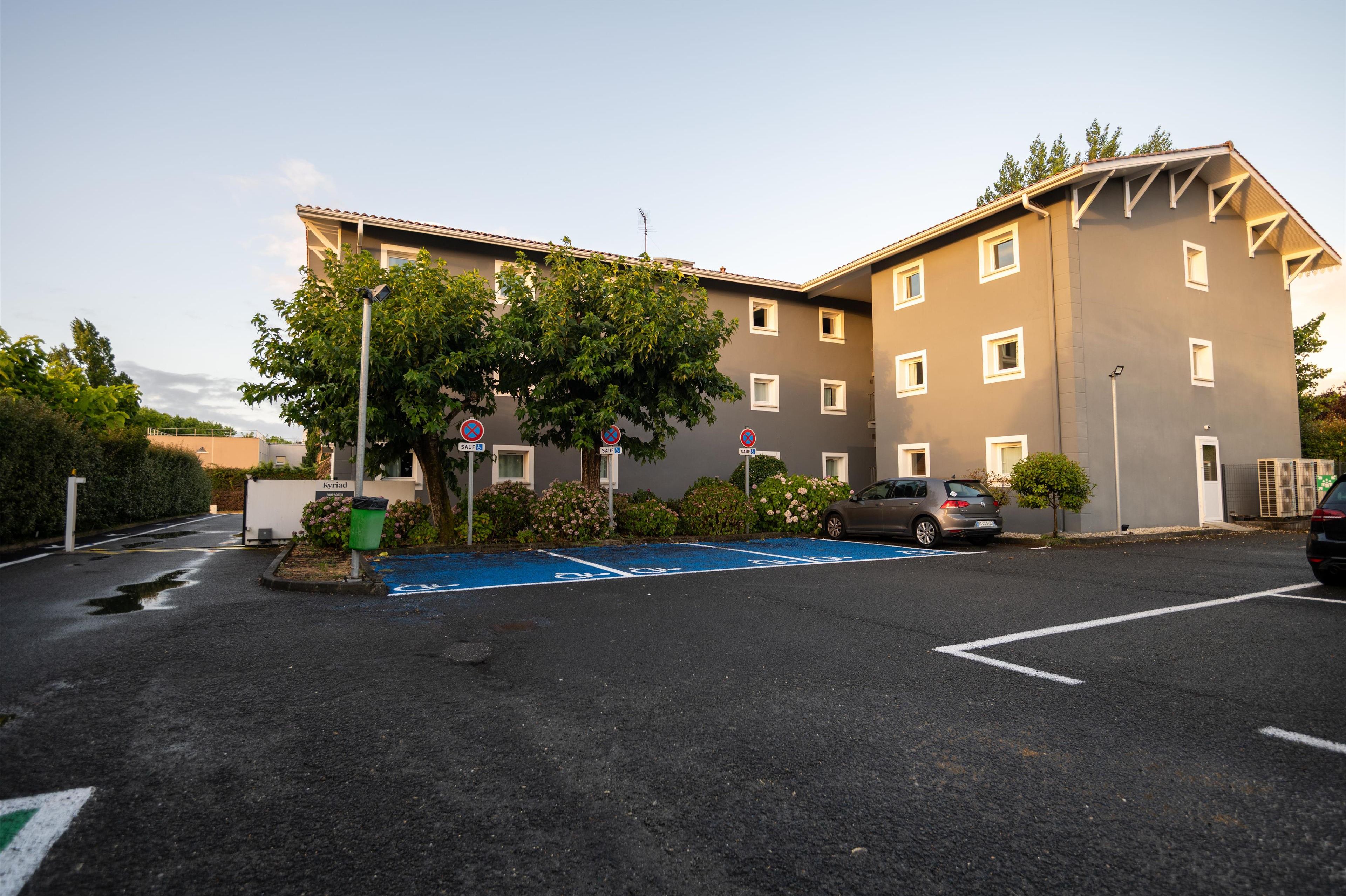 A multi-story building with a parking lot in front, featuring a blue handicapped parking space, a tree, and a small garden area.