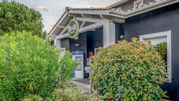 A hotel entrance with a sign reading "B&B Hotels," a reception desk, and a small garden area with various plants.