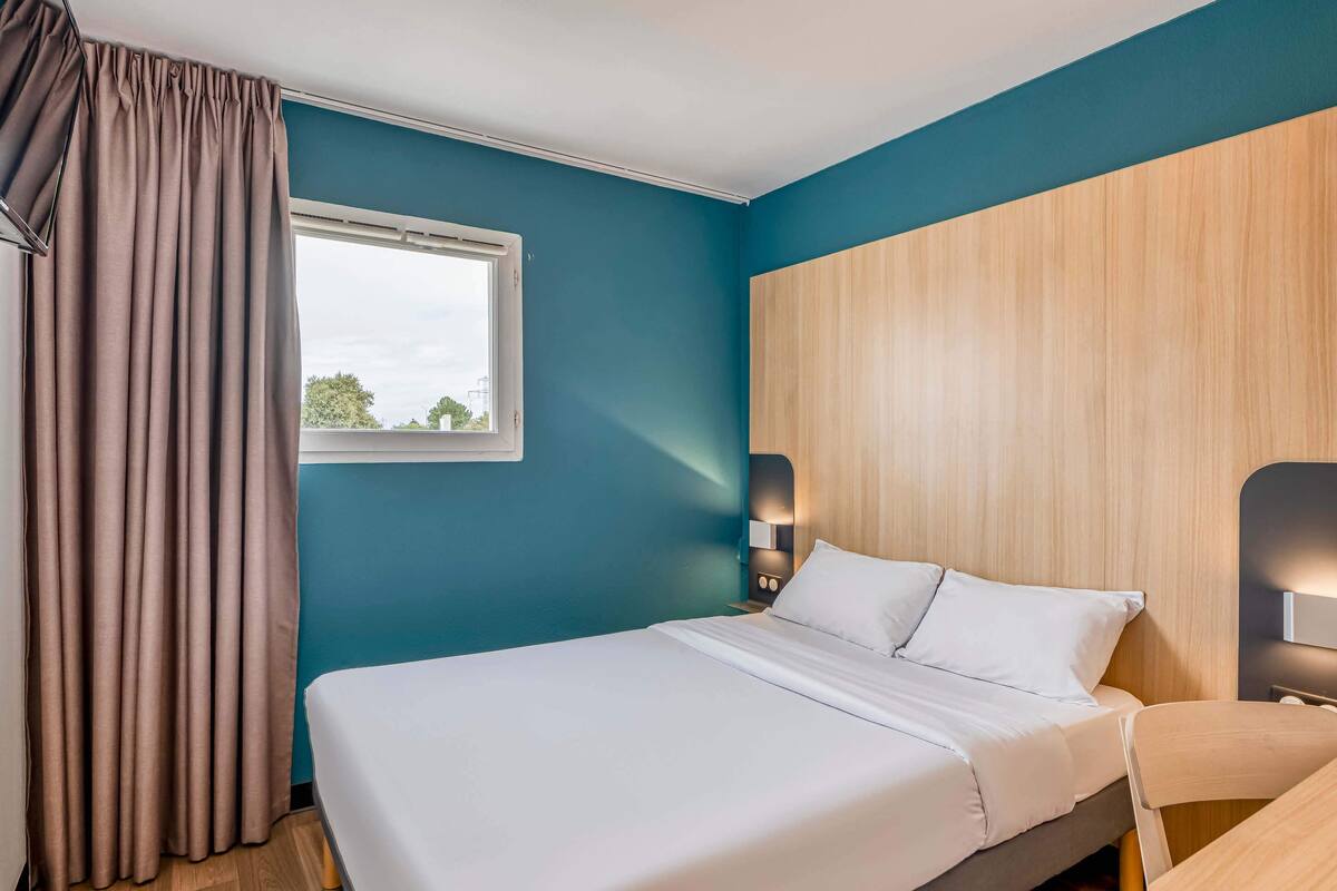 A modern hotel room with a large bed, a window with curtains, a wooden headboard, and a nightstand with a lamp.