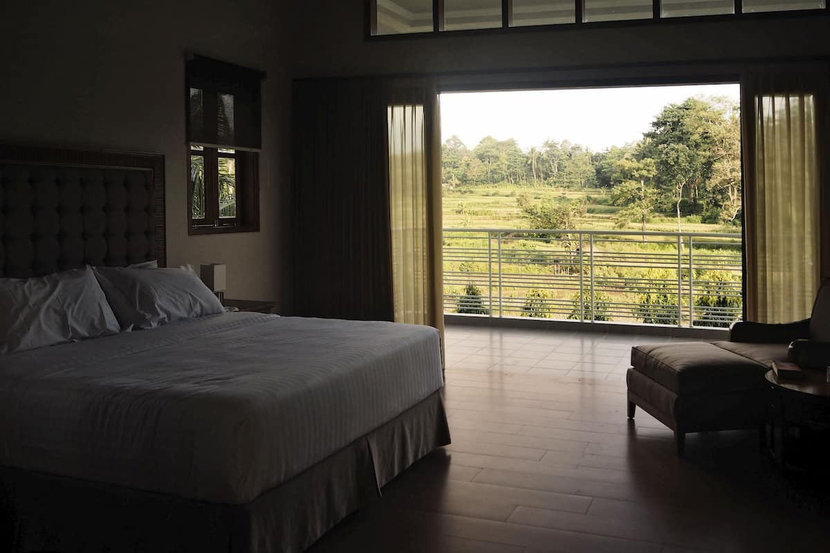 A bedroom with a large bed, a chair, and a balcony with a view of greenery.