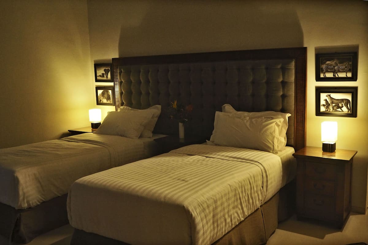 A hotel room with two single beds, a large tufted headboard, bedside tables with lamps, and framed animal pictures on the walls.