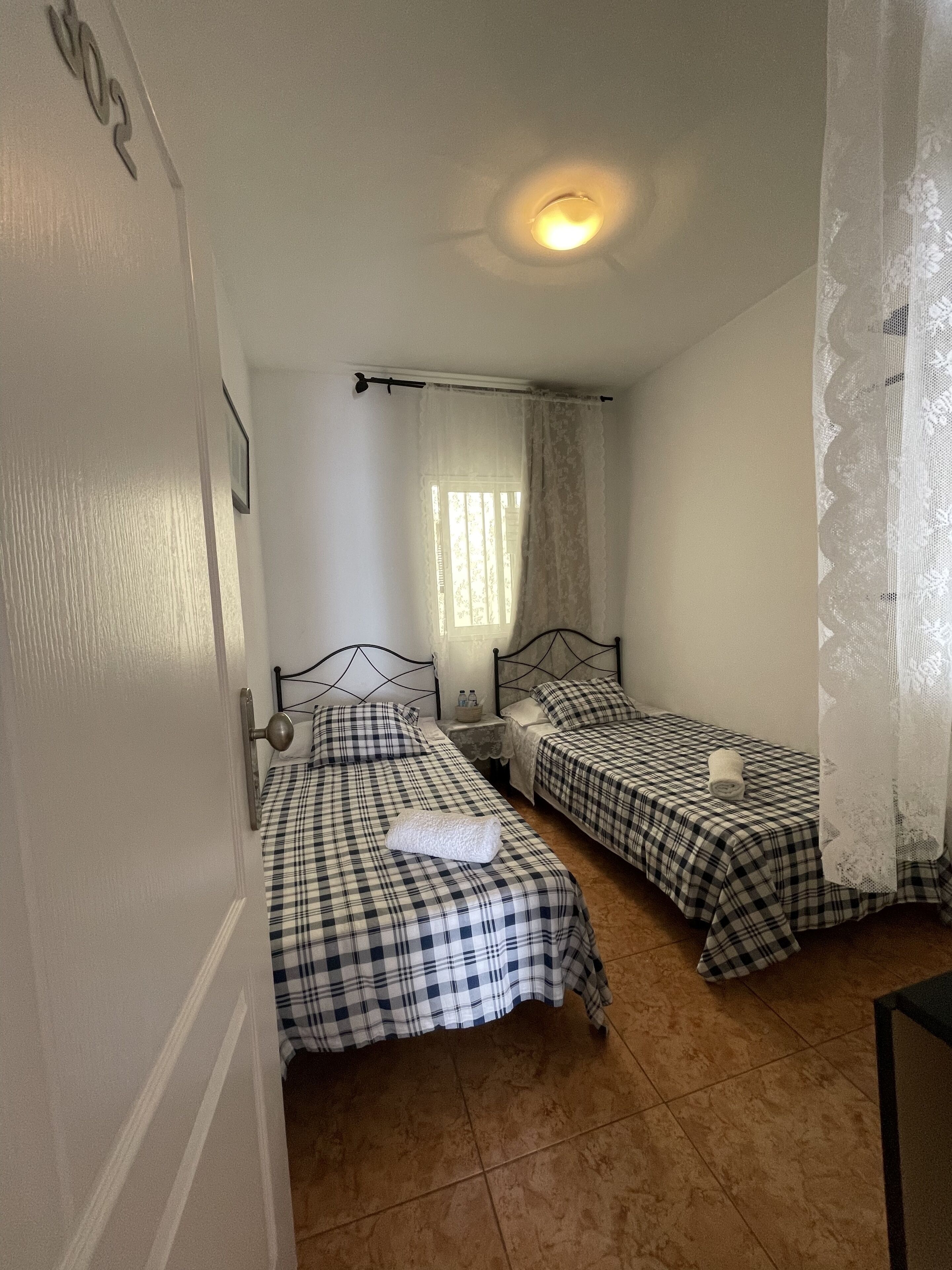 Una habitación pequeña y limpia con dos camas, una ventana con cortinas translúcidas y una lámpara en el techo.