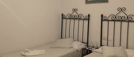 Une chambre avec deux lits simples, chacun doté d’une tête de lit en métal et d’une couvre-lit à motifs. On y trouve une petite table avec une bouteille d’eau et un tableau encadré accroché au mur, au-dessus des lits.