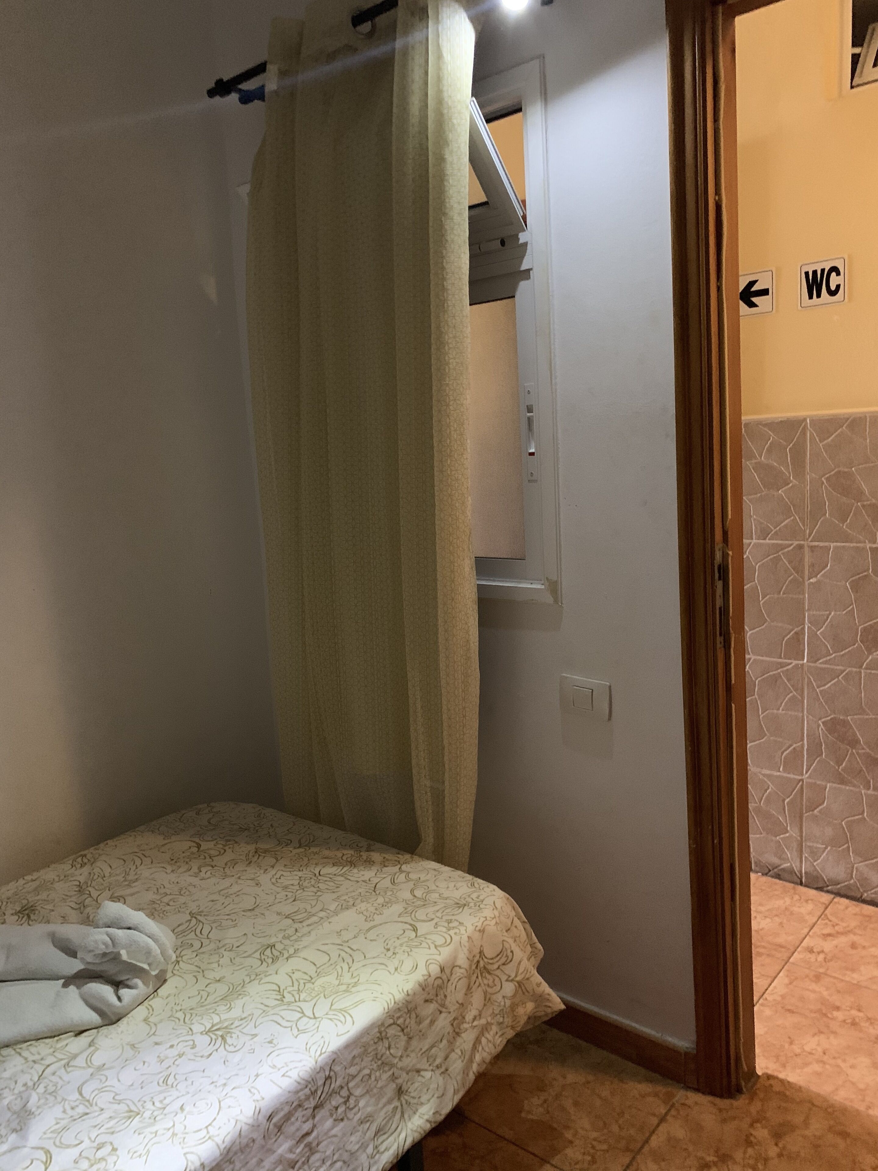 Habitación pequeña con cama individual, cortina, ventana y un cartel que indica el camino al baño.