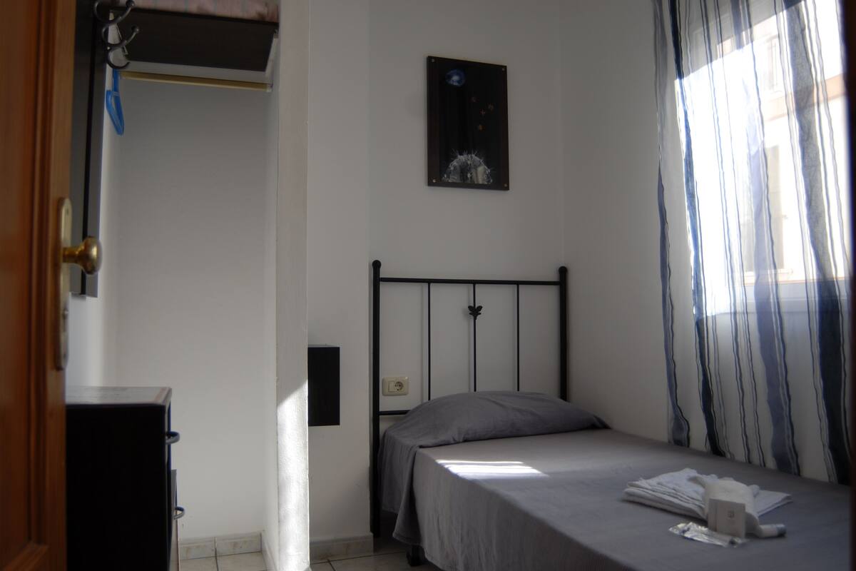 A bedroom with a bed, a bedside table, a wall-mounted light, and a window with curtains.