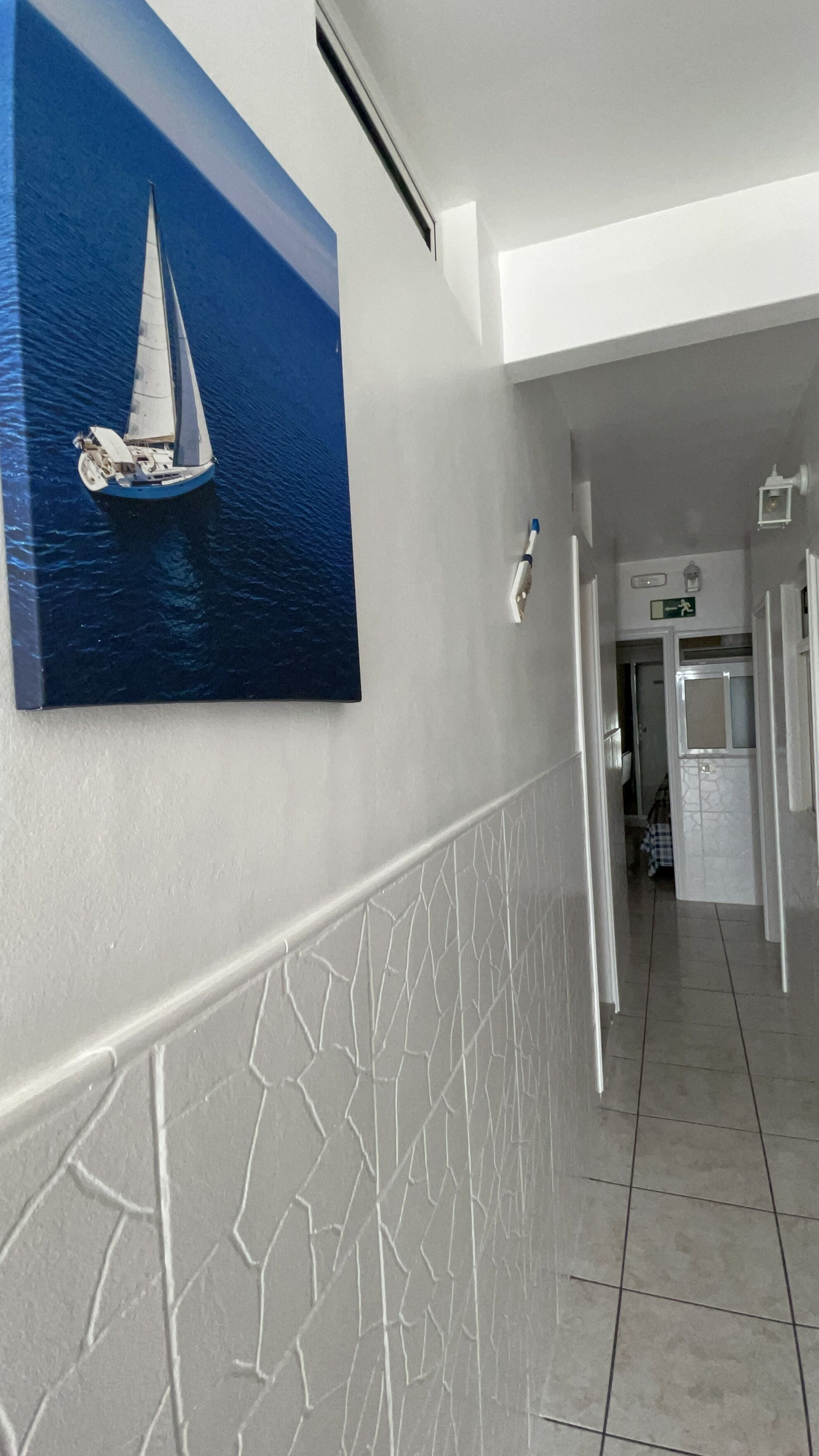 A hallway with a tiled floor, a white wall with a decorative pattern, and a painting of a sailboat on the wall.