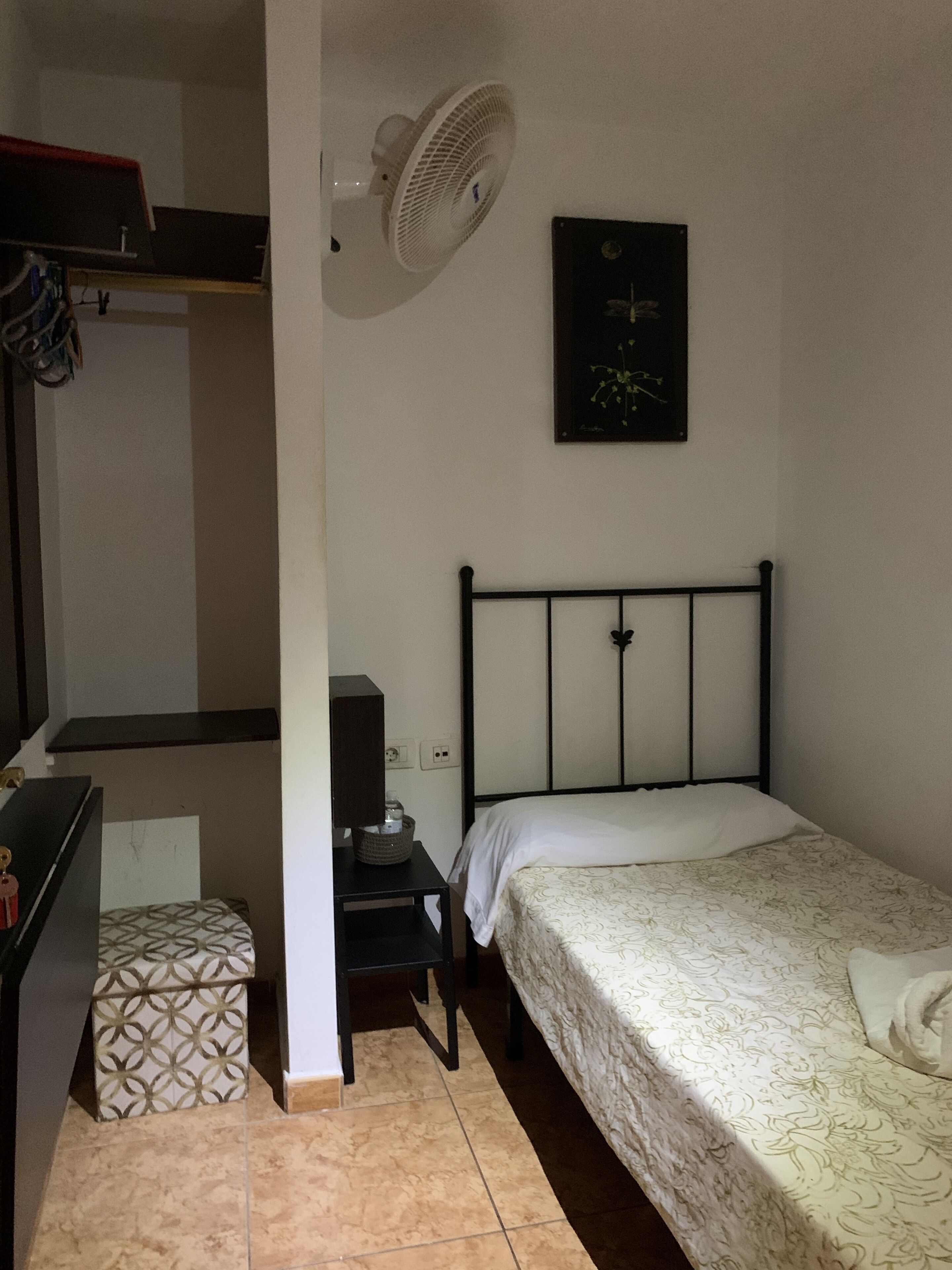 A bedroom with a bed, a nightstand, a desk, a chair, a fan, and a picture on the wall.