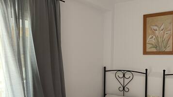 Una habitación pequeña y limpia con dos camas individuales, una mesita de noche, un cuadro en la pared y una ventana con cortinas.