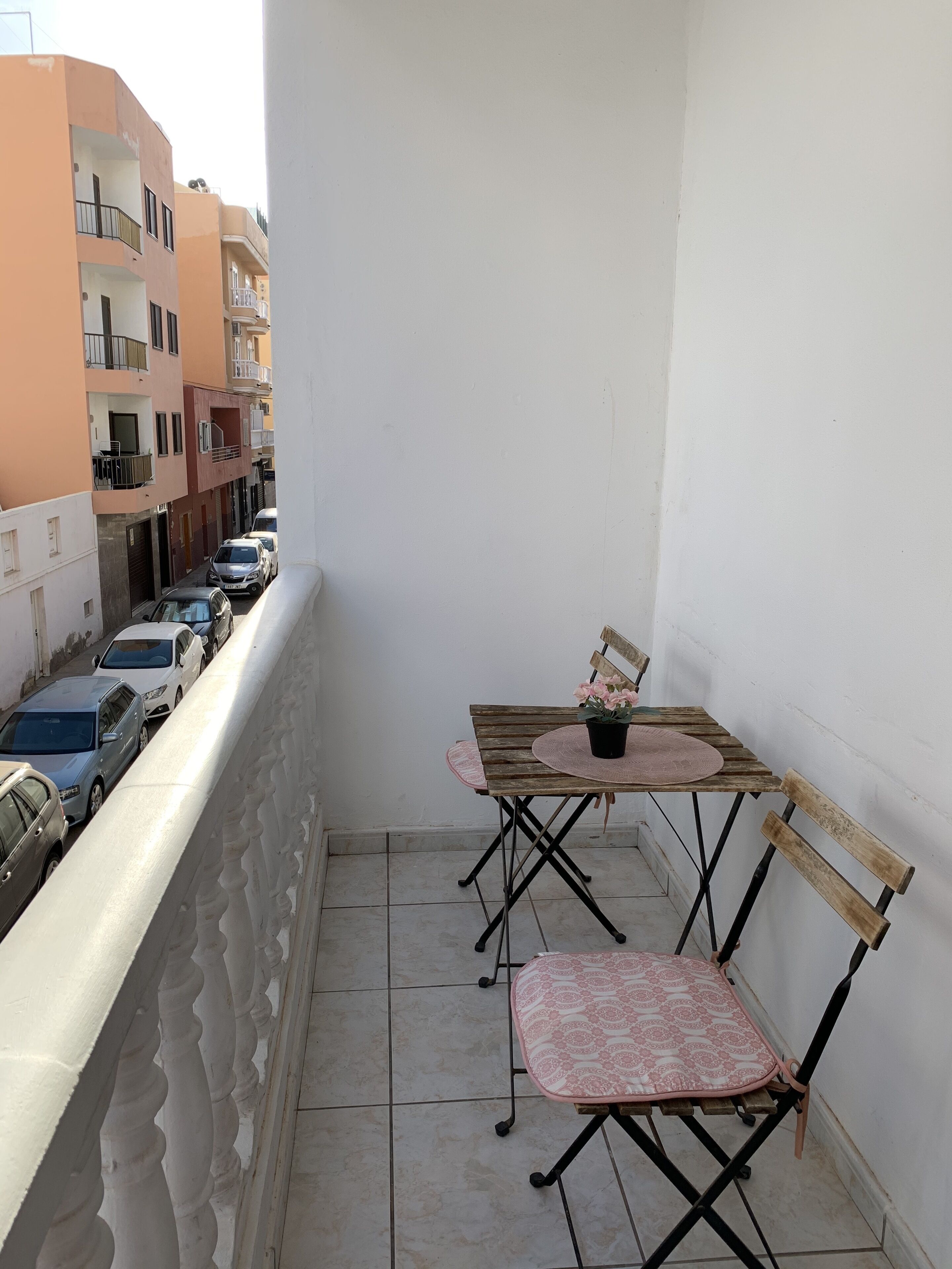 A balcony with a table and two chairs, a potted plant, and a view of parked cars and buildings.