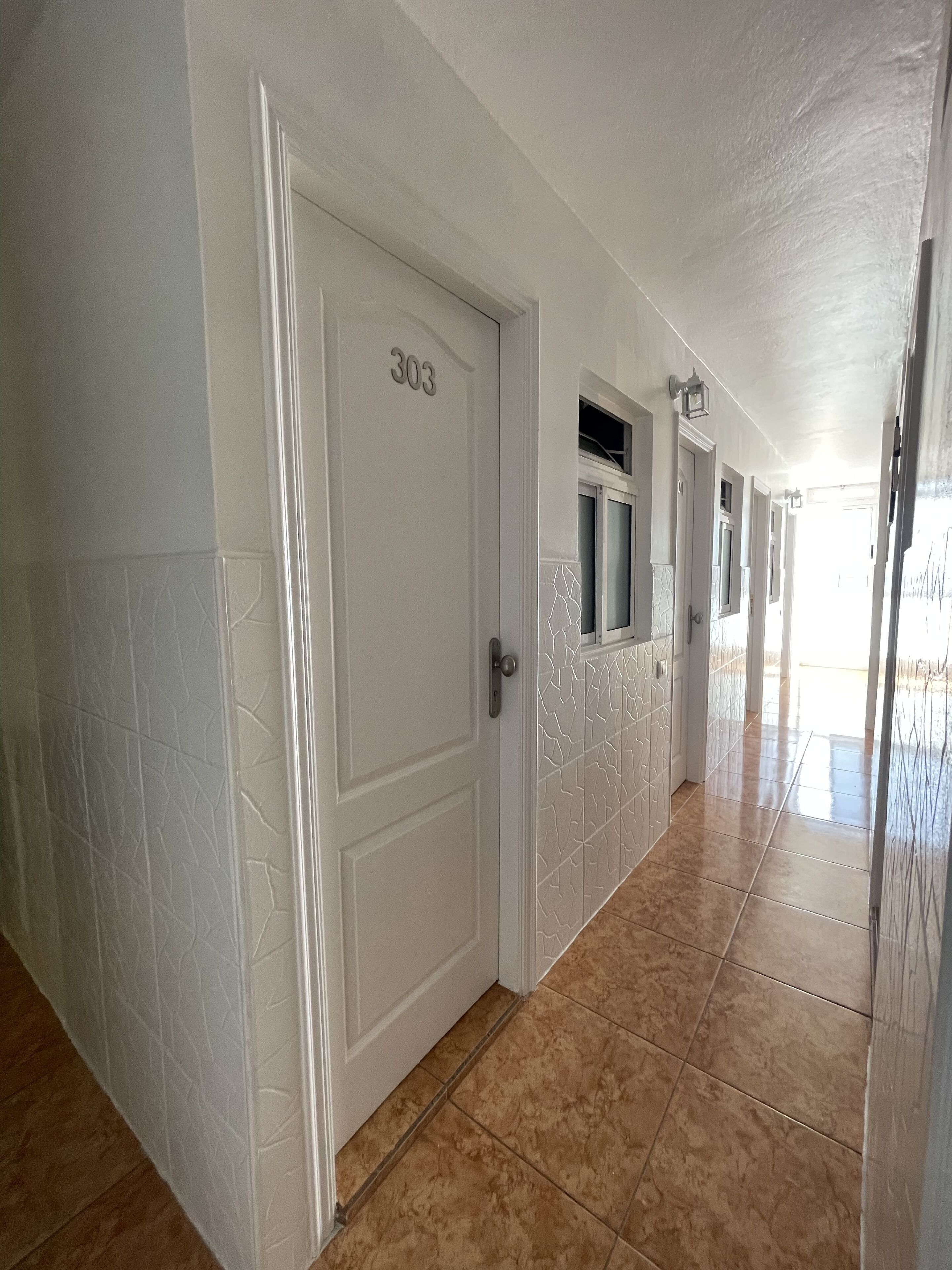 A hallway with a white door labeled 303, leading to a room with a window and a door. The hallway has patterned walls and tiled flooring.