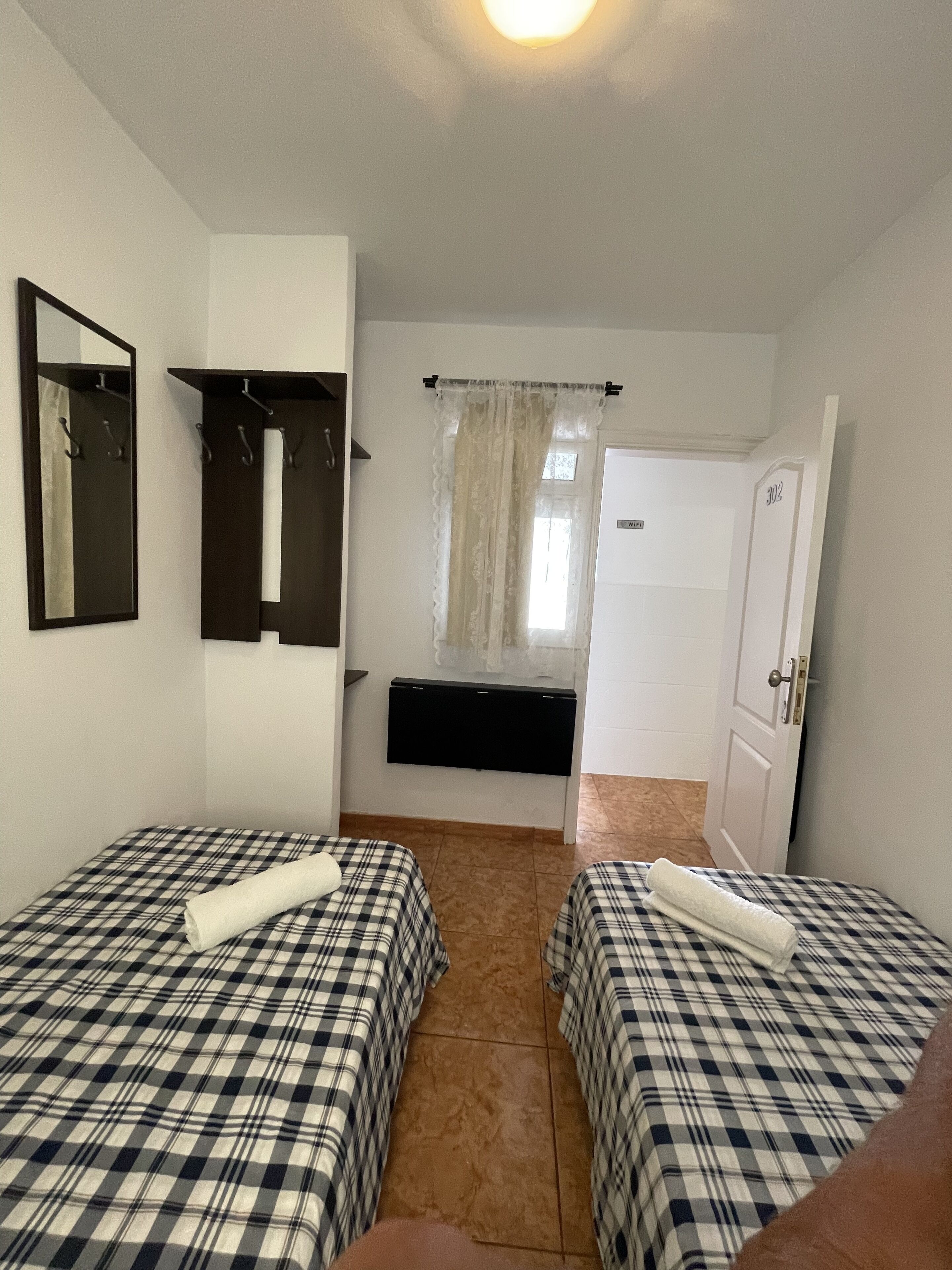 A room with two beds, a mirror, a coat rack, a door, and a window with curtains.