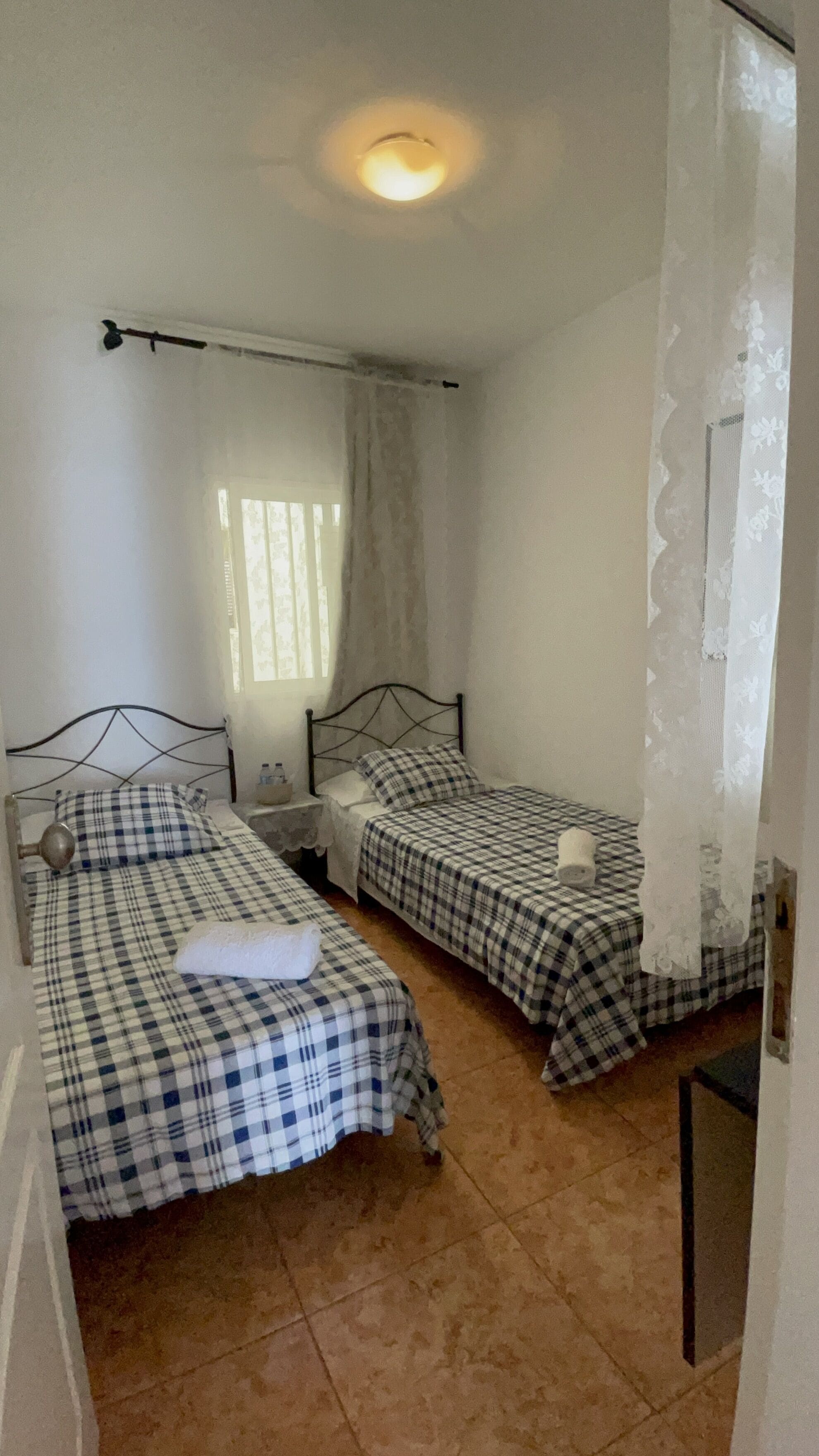Habitación con dos camas individuales, cada una con cubrecama a cuadros, una mesita de noche y una lámpara. Se ve una ventana con cortinas, una lámpara en el techo y una puerta con cortina floral.