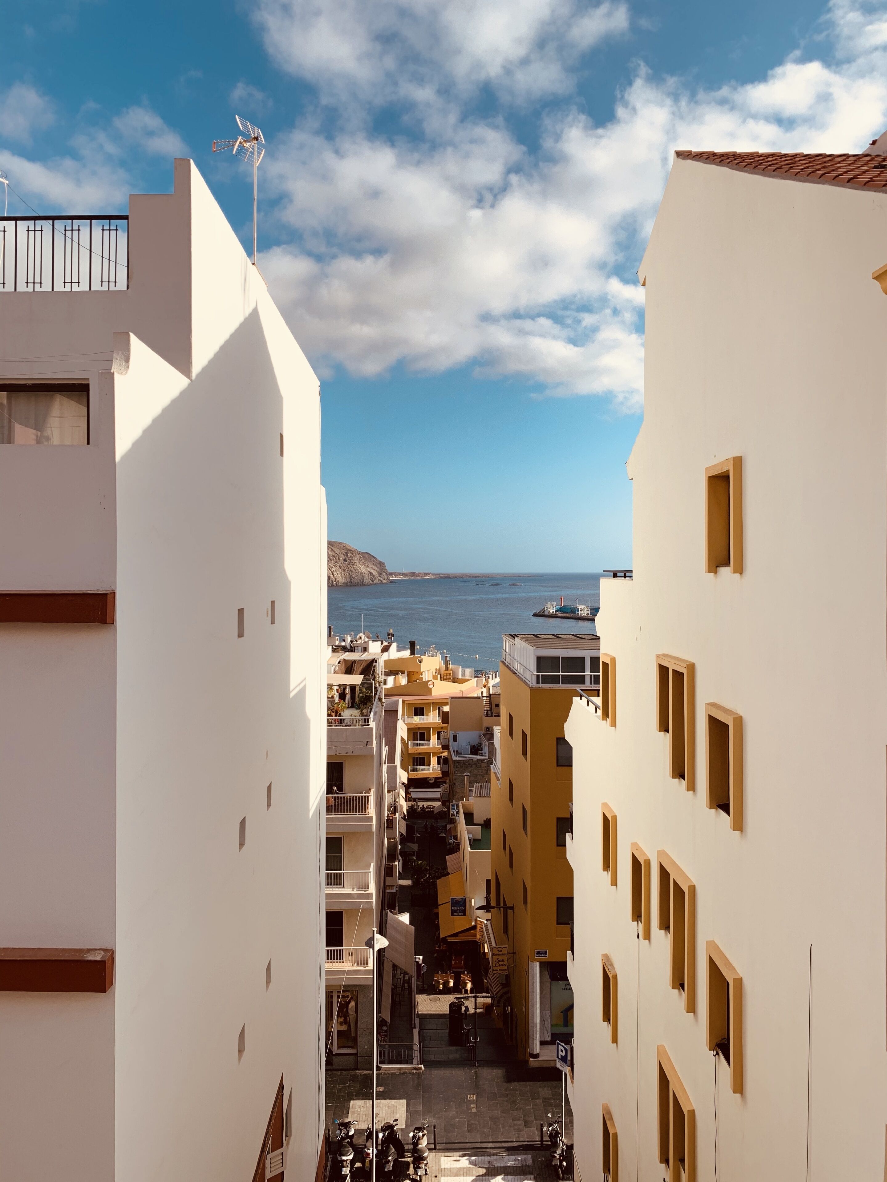 A narrow street with buildings on either side, leading to a coastal view.