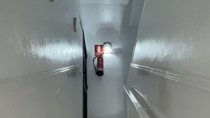 Uma escadaria com degraus e corrimãos brancos, que leva a um patamar com um extintor e uma luminária.