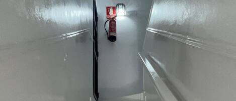 Una escalera con escalones y barandas blancas que conduce a una plataforma con un extintor y una lámpara.