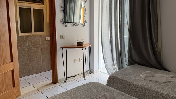 Quarto de hotel com duas camas, uma mesinha com uma cesta, um espelho e uma janela com cortinas.