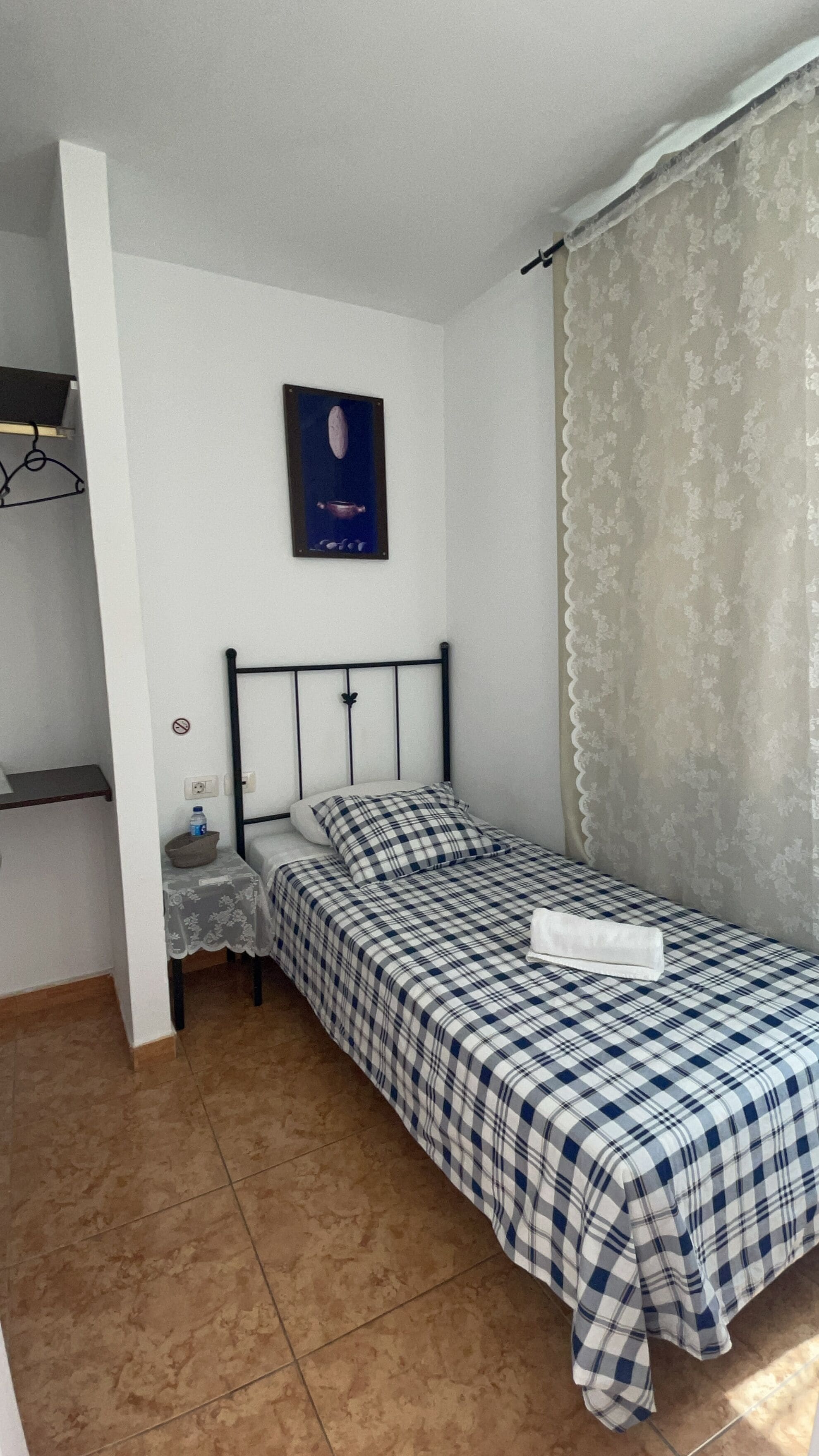 Une petite chambre avec un lit simple, recouverte d’une courtepointe à carreaux, une table de chevet avec une bouteille d’eau, un tableau accroché au mur et une fenêtre ornée d’un rideau léger.