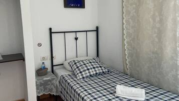 Een kleine eenpersoonskamer met een geruit sprei, een nachtkastje met een fles water, een aan de muur hangend schilderij en een raam met een dunne gordijn.