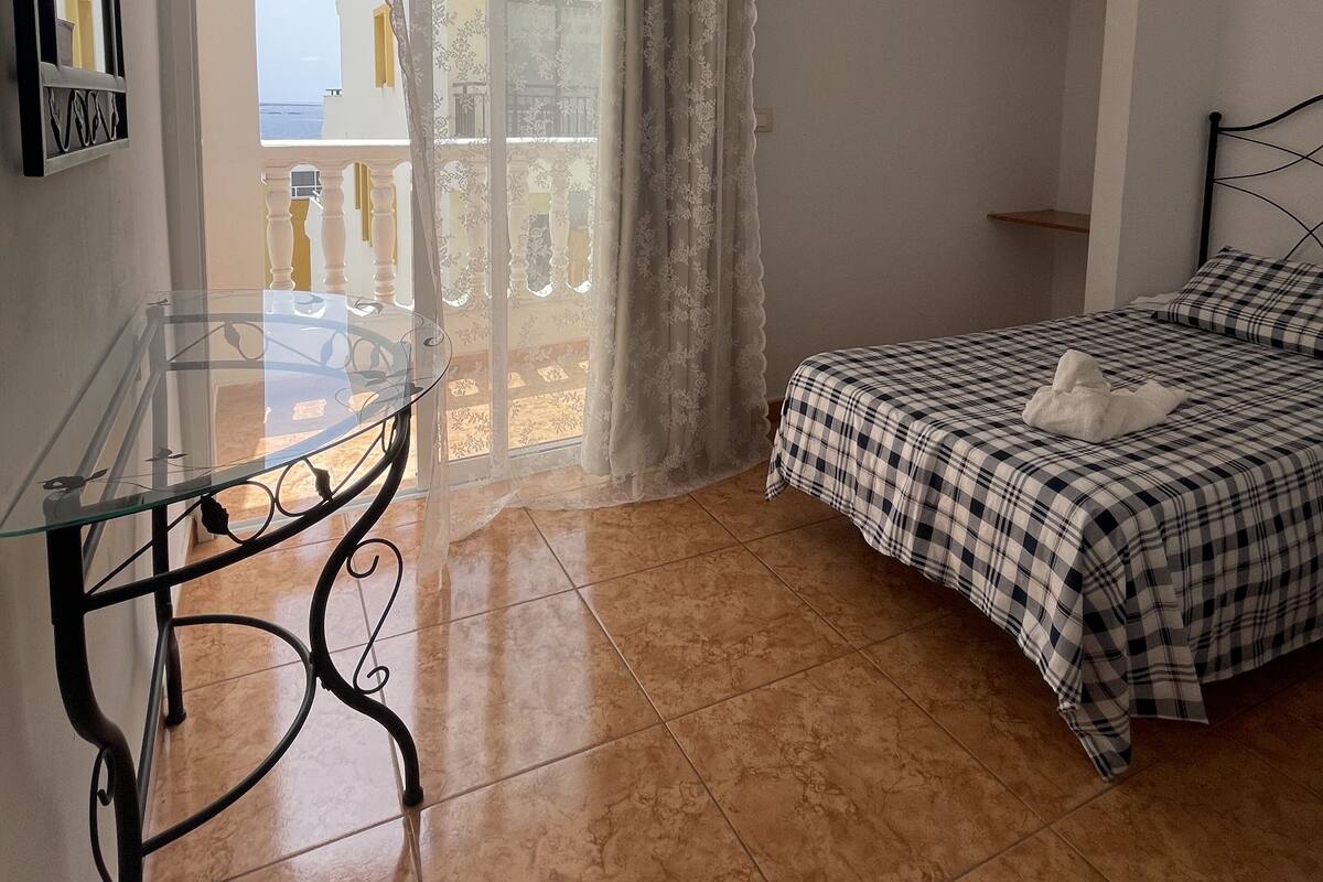 A bedroom with a glass table, a bed with checkered bedding, and a balcony with a view.