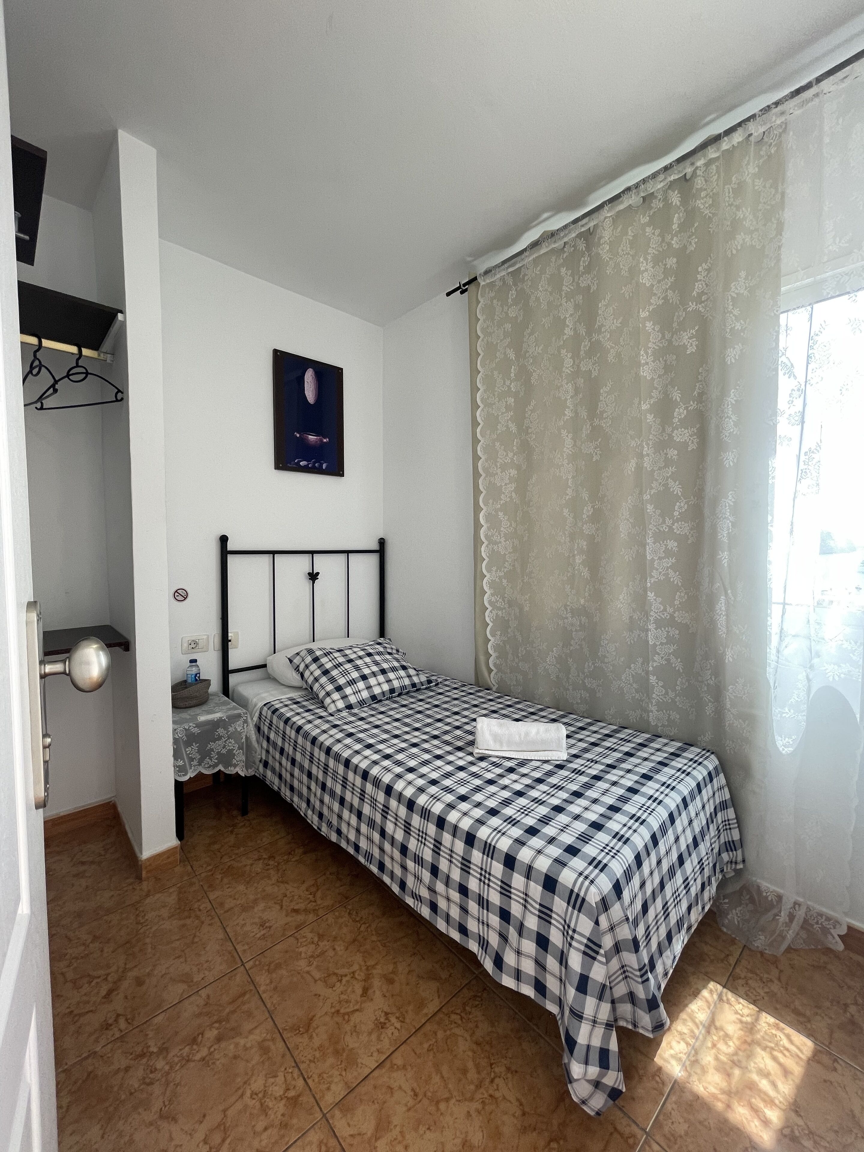 A single bed with a checkered bedspread, a nightstand with a lamp, a wall-mounted picture, and a window with sheer curtains.